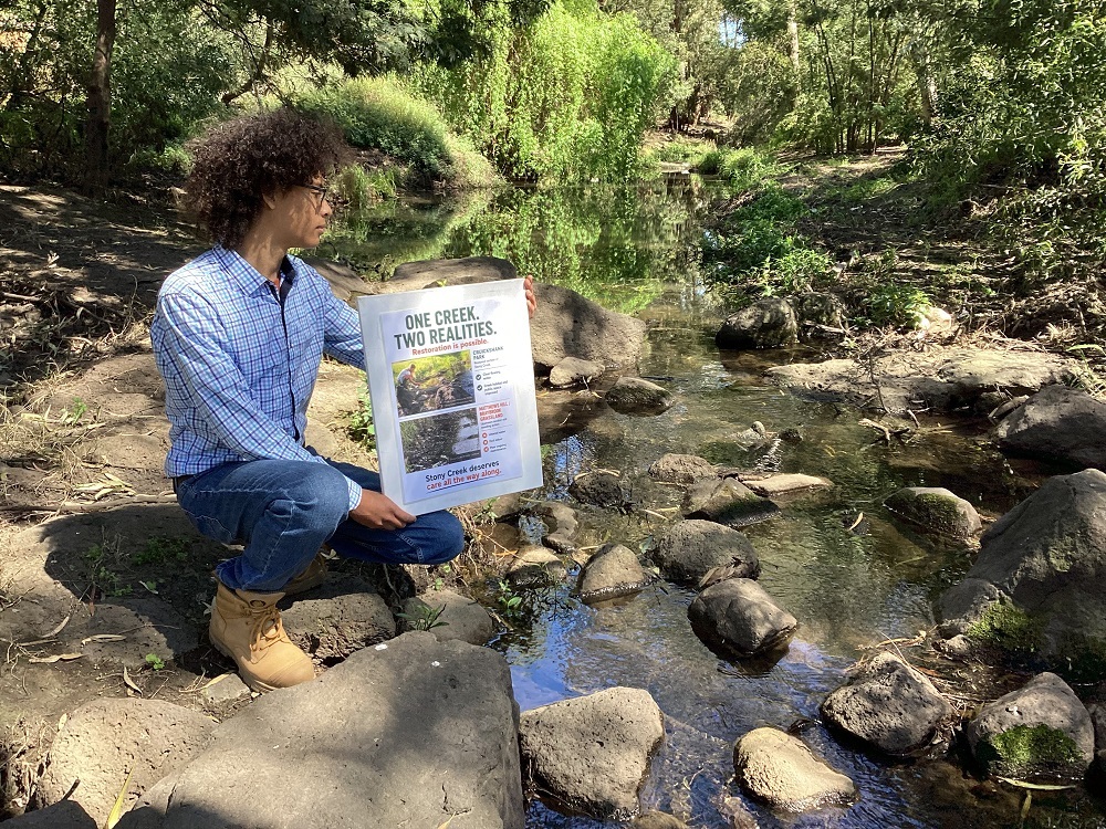 Chris holding the One Creek Two Realities poster beside Stony Creek at Cruickshank Park